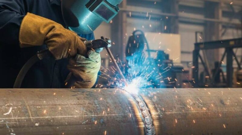 Welder sedang mengelas menggunakan teknik MIG dan TIG dengan helm dan sarung tangan melatih pola sambungan kuat rapi.